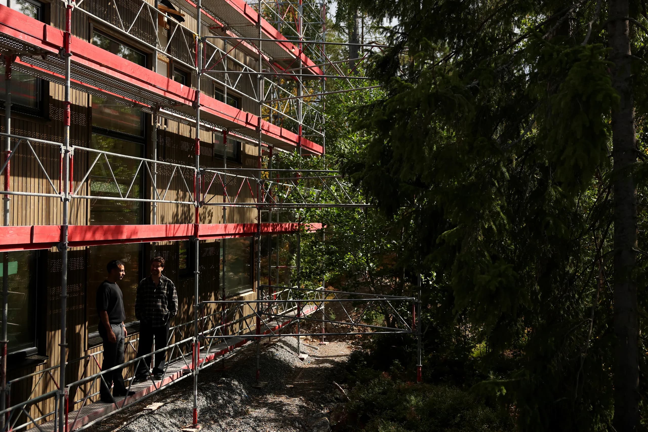 Två medarbetare på byggnadsställningen längs husets träfasad i tallskogen